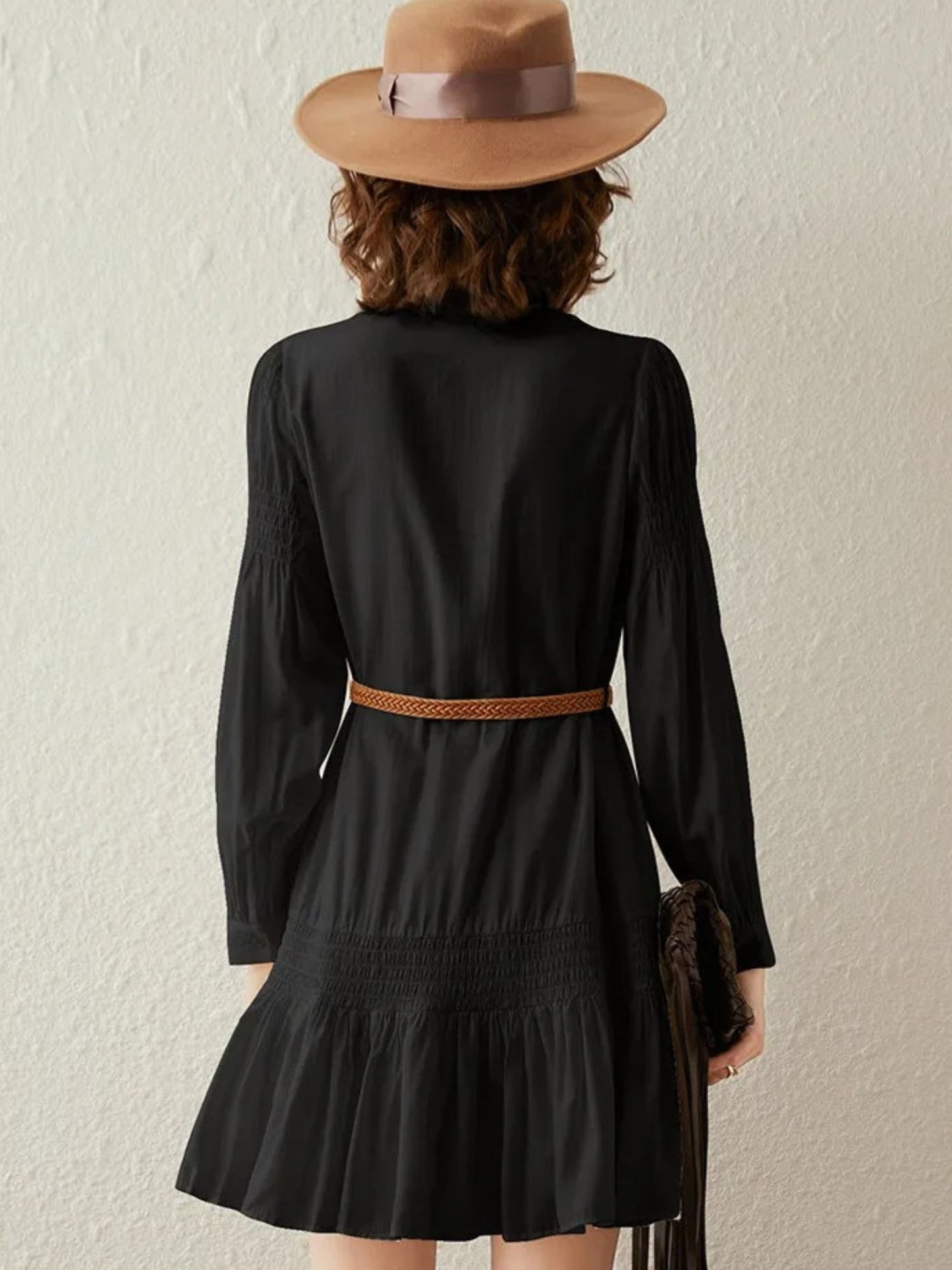 Back view of a woman wearing a black boho-style long sleeve dress with a tie detail in the neckline and a brown belt