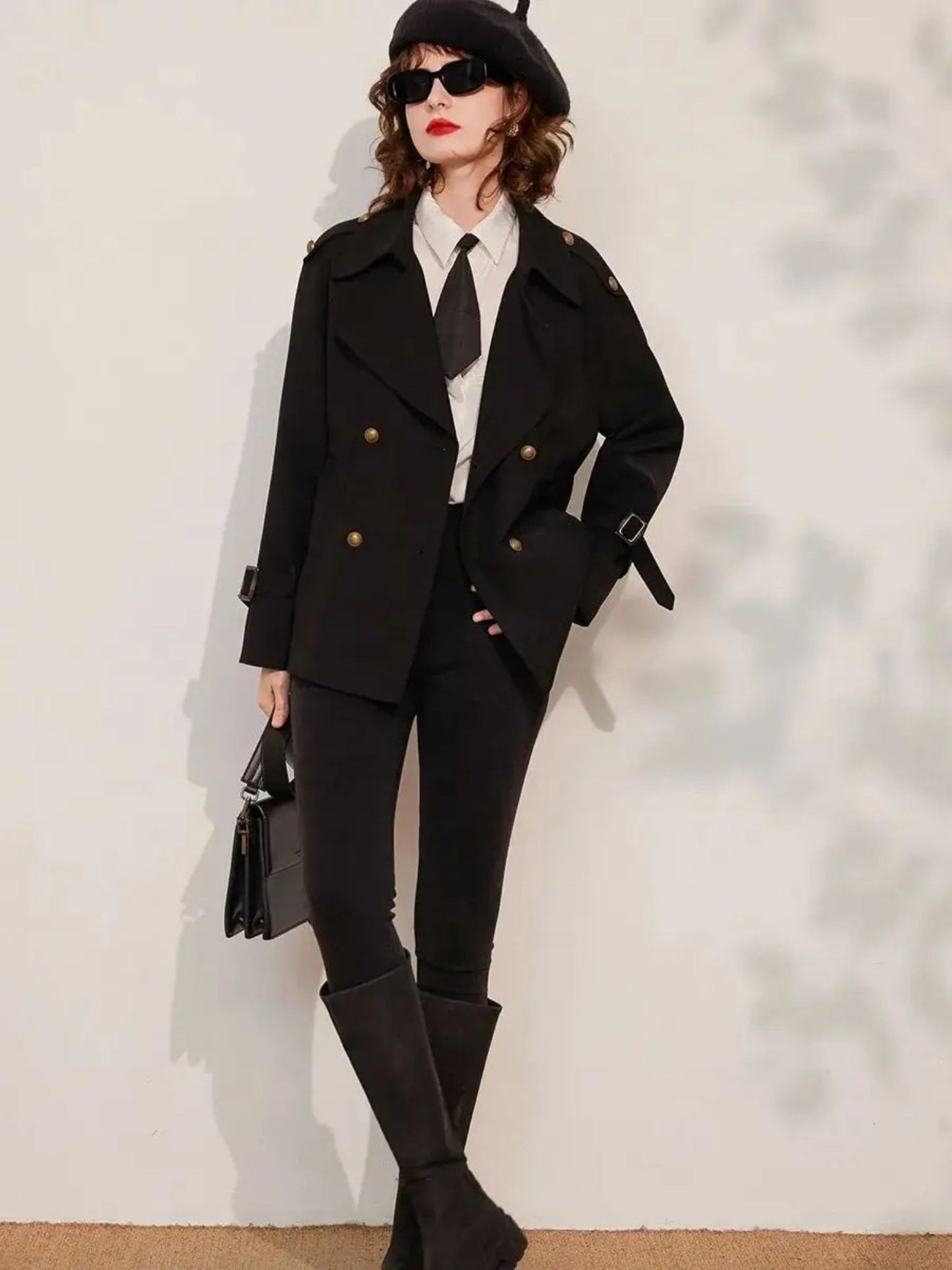 Woman leaning against wall wearing black blazer with gold buttons, white shirt, tie, and black trousers