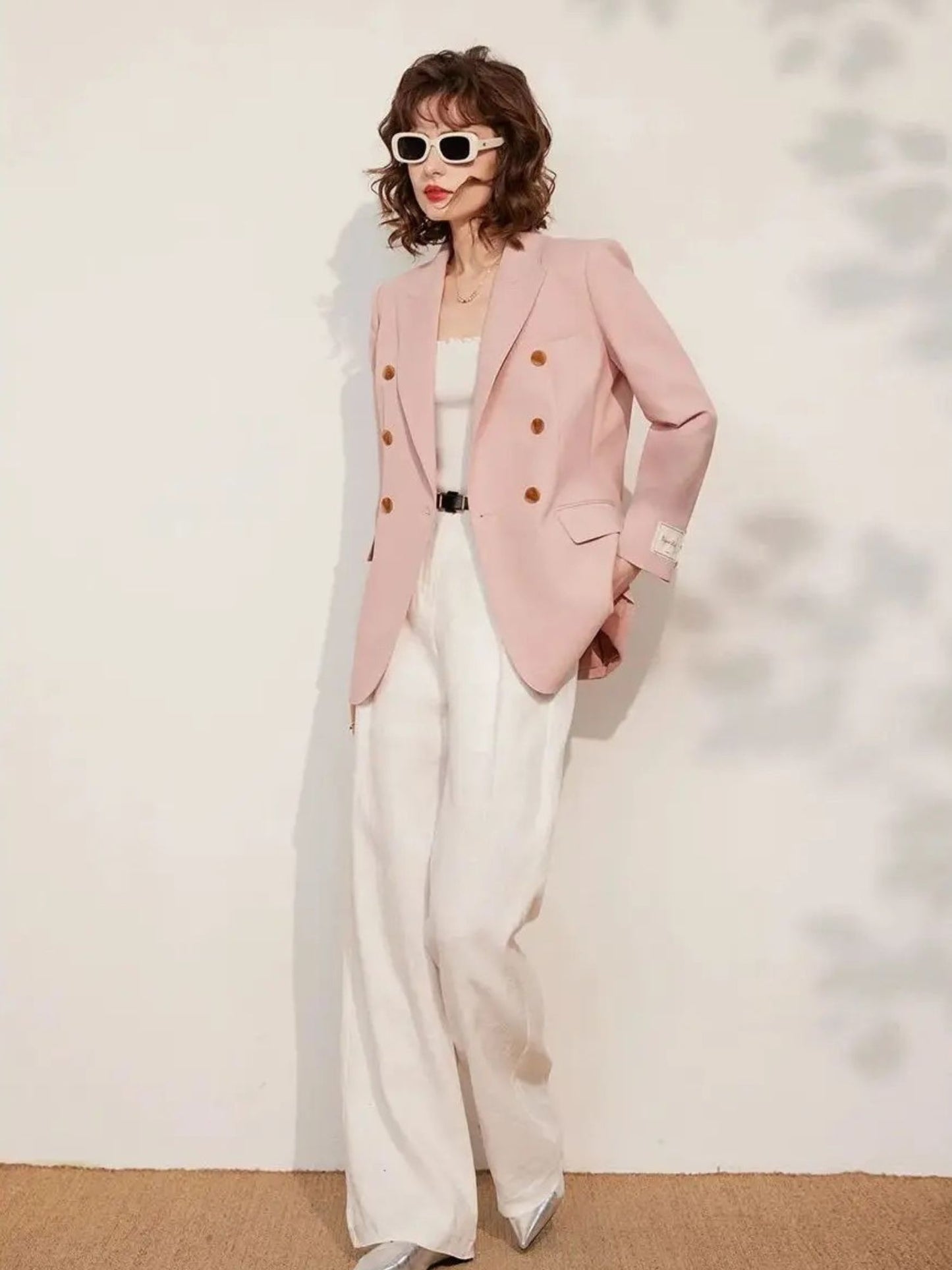 Full-body view of the woman standing against a light background, wearing the pink blazer and white wide-leg trousers, styled with sunglasses and heels